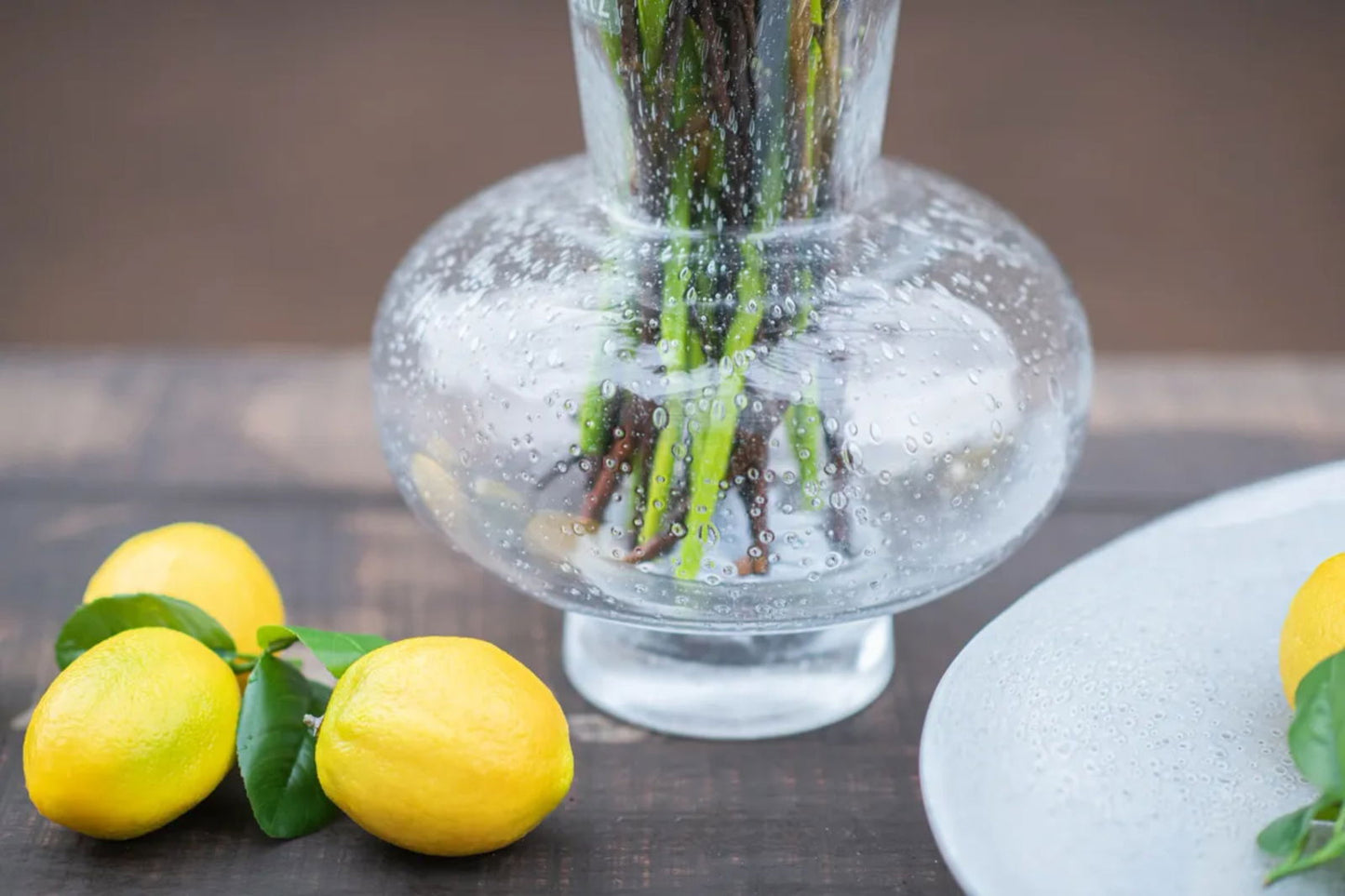 Clear Lowlands Design vase with textured bubbles holding flower stems on wooden table with lemons nearby