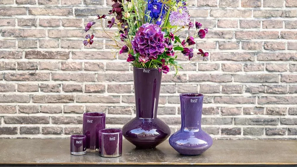 Lowlands Design plum glass vase set with tall floral arrangement and matching smaller vase and candle holders on wooden shelf