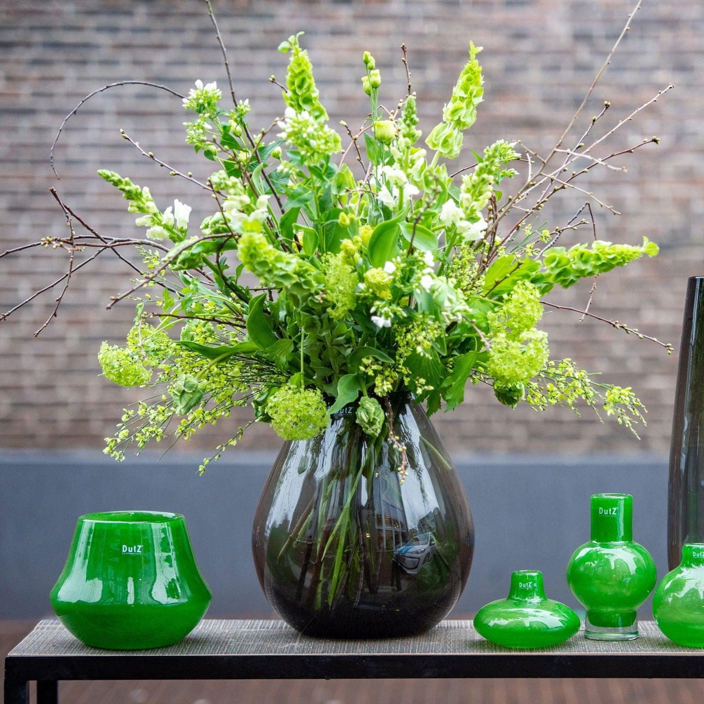 Lowlands Design glass vase with green floral arrangement on black table with green decorative items