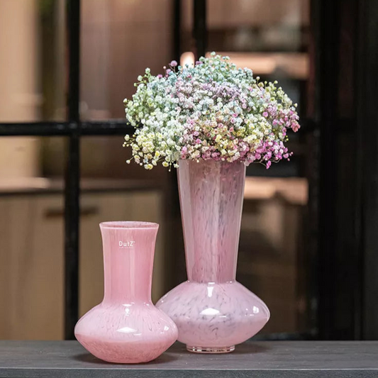 Two pink vases with a bouquet of flowers on a table against a brick wall.